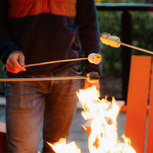 Guest roasting marshmallows over a fire pit at Riverhouse Lodge with signature orange chairs and Deschutes River views in the background during Stars & S’mores event.