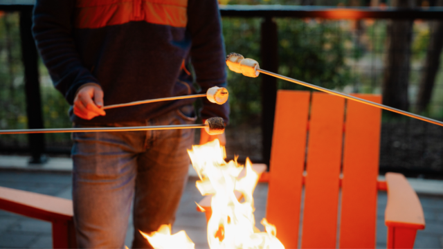 Guest roasting marshmallows over a fire pit at Riverhouse Lodge with signature orange chairs and Deschutes River views in the background during Stars & S’mores event.