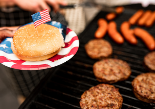 A grill with burgers and sausages, a plate with a bun and a small flag, suggesting a festive barbecue gathering.