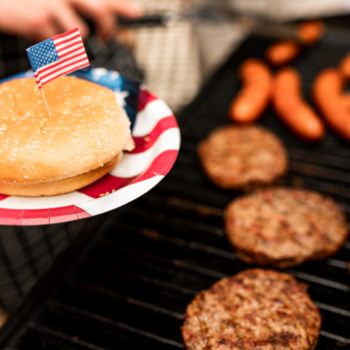 A grill with burgers and sausages, a plate with a bun and a small flag, suggesting a festive barbecue gathering.