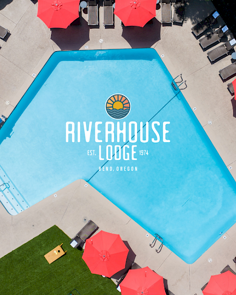 An aerial view of a swimming pool at Riverhouse Lodge, surrounded by red umbrellas, lounge chairs, and a lush green area in Bend, Oregon.