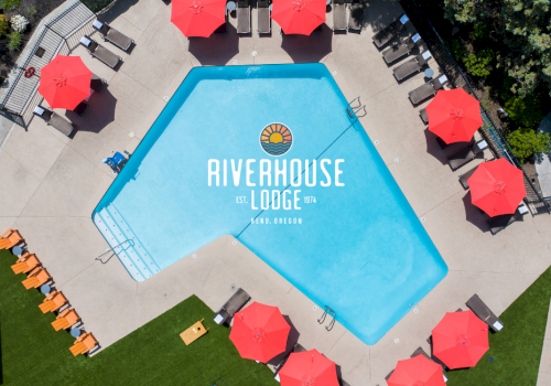 An aerial view of a swimming pool at Riverhouse Lodge, surrounded by red umbrellas, lounge chairs, and a lush green area in Bend, Oregon.