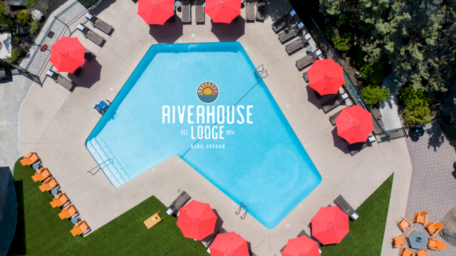 An aerial view of a swimming pool at Riverhouse Lodge, surrounded by red umbrellas, lounge chairs, and a lush green area in Bend, Oregon.