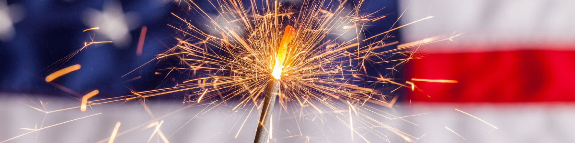 A burning sparkler in focus with an American flag in the blurred background.