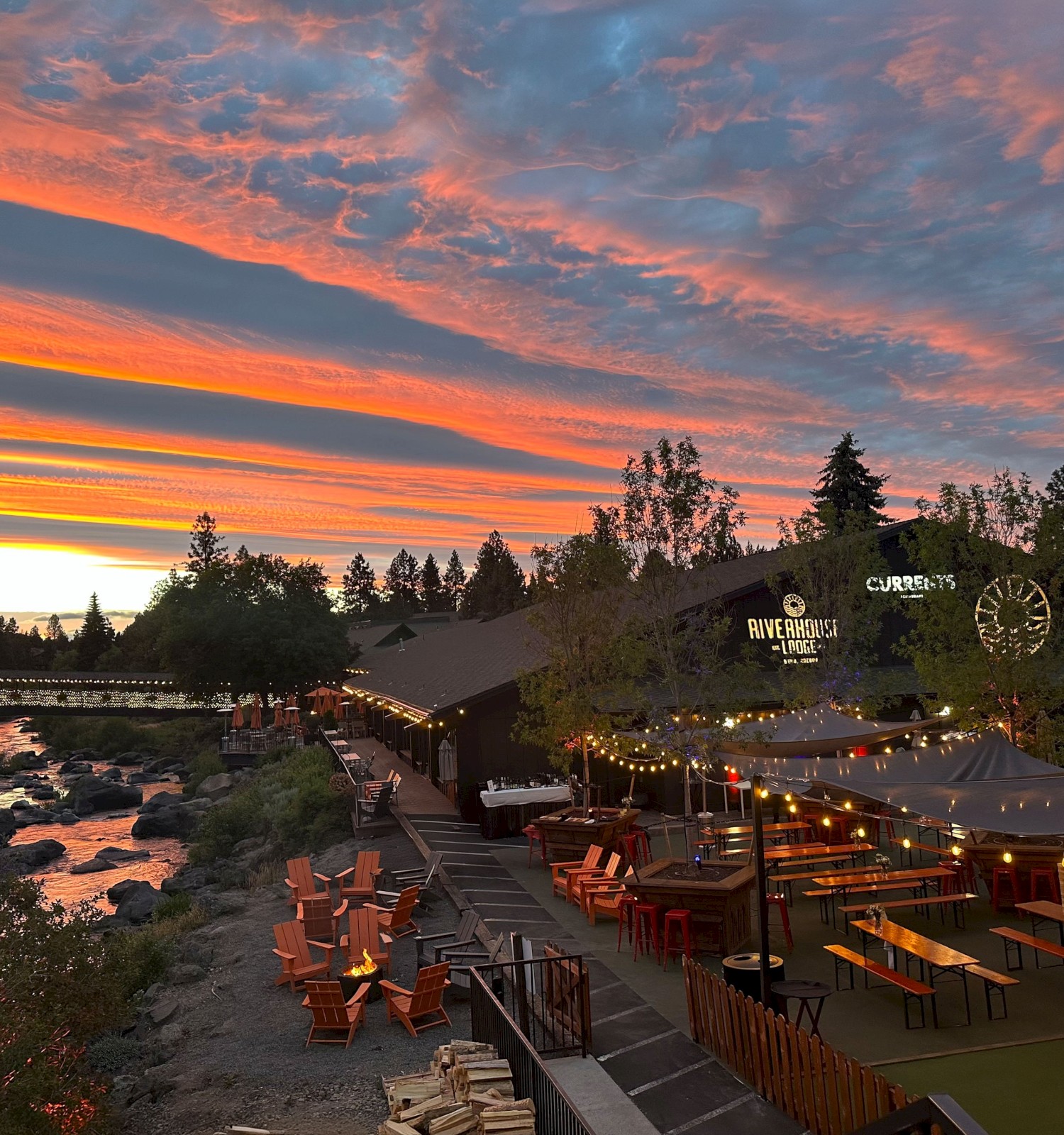 An outdoor dining area by a river at Riverhouse Lodge in Bend, Oregon, with string lights and seating, under a dramatic sunset sky with vibrant clouds and trees around.