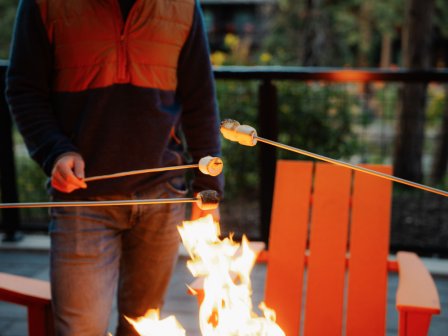 A person is roasting marshmallows over an outdoor fire with orange chairs in the background at Riverhouse Lodge in Bend, Oregon