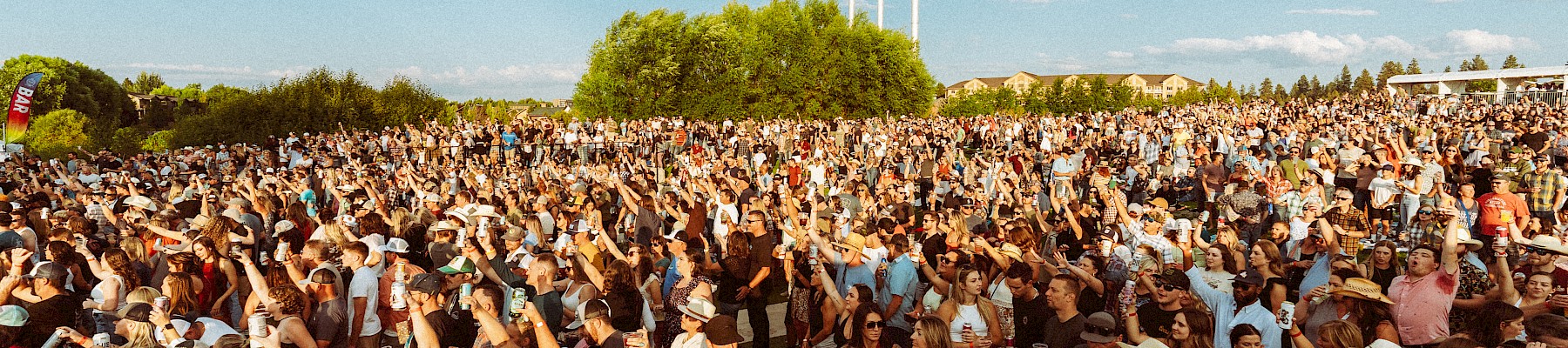 A large crowd gathered outdoors at Hayden Homes Amphitheater in Bend, Oregon, enjoying an event under a clear blue sky with scattered clouds, surrounded by green trees.