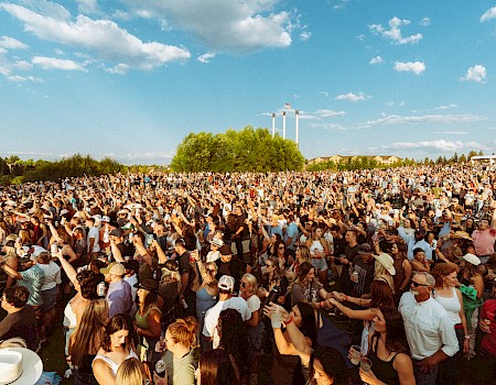 A large crowd gathered outdoors at Hayden Homes Amphitheater in Bend, Oregon, enjoying an event under a clear blue sky with scattered clouds, surrounded by green trees.