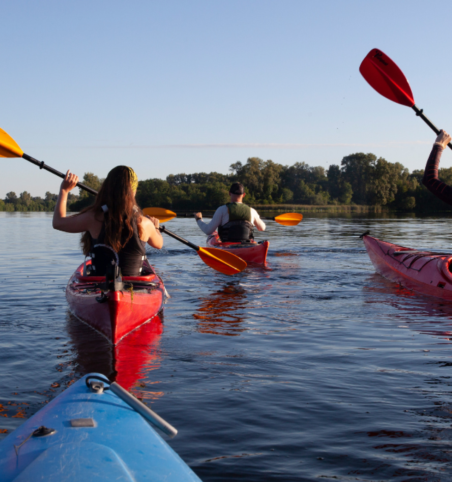 People are kayaking on the Deschutes River in Bend, Oregon with kayaks rented at Riverhouse Lodge, with trees lining the distant bank, under a clear blue sky.