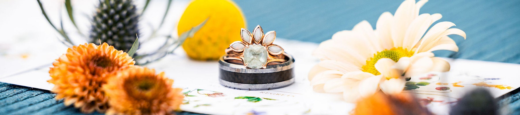 Rings with a floral design are placed on a colorful surface, surrounded by various flowers and natural elements.