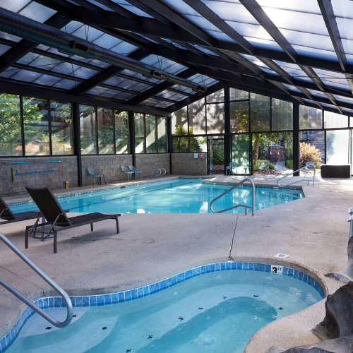 Spacious indoor pool and hot tub area at Riverhouse Lodge with lounge chairs and floor-to-ceiling windows overlooking the Deschutes River greenery.
