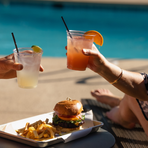 Two people clink drinks by a pool, with a tray holding a burger and fries in the foreground, sunlit and relaxed atmosphere.