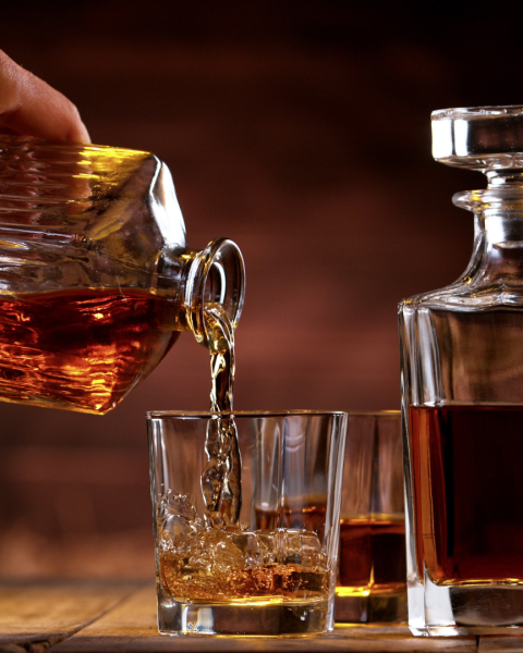 A hand pours whiskey into a glass from a decanter, with another filled glass and a decanter beside it on a wooden table.