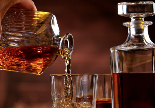 A hand pours whiskey into a glass from a decanter, with another filled glass and a decanter beside it on a wooden table.