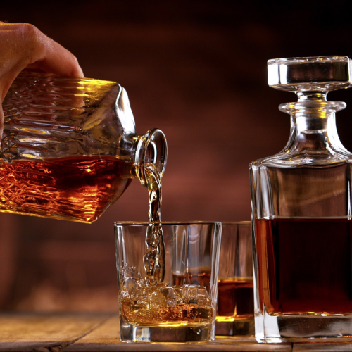 A hand pours whiskey into a glass from a decanter, with another filled glass and a decanter beside it on a wooden table.
