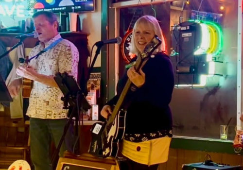Big Treble is performing in a bar, with one person singing and playing guitar, another on bass, and colorful neon lights in the background. Live music at Riverhouse Lodge in Bend Oregon