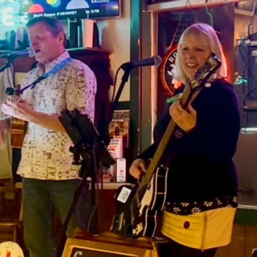 Big Treble is performing in a bar, with one person singing and playing guitar, another on bass, and colorful neon lights in the background. Live music at Riverhouse Lodge in Bend Oregon
