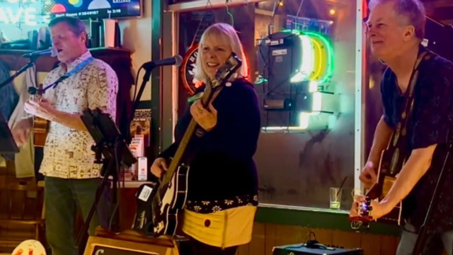 Big Treble is performing in a bar, with one person singing and playing guitar, another on bass, and colorful neon lights in the background. Live music at Riverhouse Lodge in Bend Oregon
