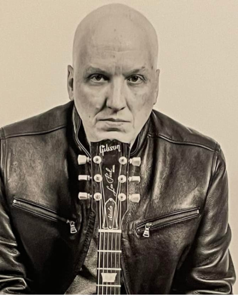 Musician David Miller in a leather jacket is seated with a guitar in front, appearing in a serious pose against a plain background. Free live show at Riverhouse Lodge in Bend Oregon 08/22/25