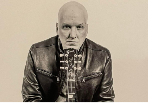 Musician David Miller in a leather jacket is seated with a guitar in front, appearing in a serious pose against a plain background. Free live show at Riverhouse Lodge in Bend Oregon 08/22/25