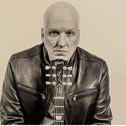 Musician David Miller in a leather jacket is seated with a guitar in front, appearing in a serious pose against a plain background. Free live show at Riverhouse Lodge in Bend Oregon 08/22/25