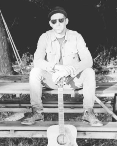 Kevin Conness of Bim Jeam & The Sadness sits on a picnic table outdoors, wearing a jacket and sunglasses, with a guitar standing upright in front of them. Free live show at Riverhouse Lodge in Bend Oregon 08/29/25
