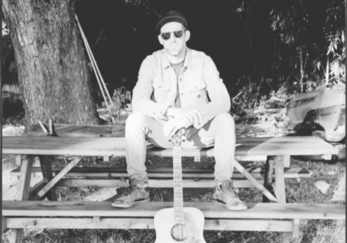 Kevin Conness of Bim Jeam & The Sadness sits on a picnic table outdoors, wearing a jacket and sunglasses, with a guitar standing upright in front of them. Free live show at Riverhouse Lodge in Bend Oregon 08/29/25