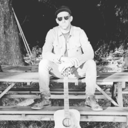 Kevin Conness of Bim Jeam & The Sadness sits on a picnic table outdoors, wearing a jacket and sunglasses, with a guitar standing upright in front of them. Free live show at Riverhouse Lodge in Bend Oregon 08/29/25