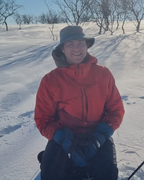Ian Lafky in a red jacket and hat sits on snowy terrain with ski poles, surrounded by bare trees under a clear blue sky. Free live music at Riverhouse Lodge in Bend Oregon August 3