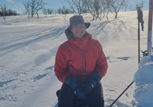 Ian Lafky in a red jacket and hat sits on snowy terrain with ski poles, surrounded by bare trees under a clear blue sky. Free live music at Riverhouse Lodge in Bend Oregon August 3
