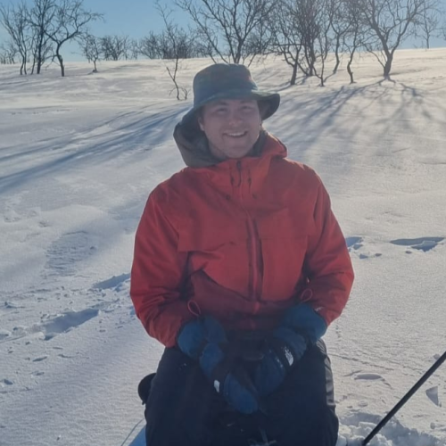 Ian Lafky in a red jacket and hat sits on snowy terrain with ski poles, surrounded by bare trees under a clear blue sky. Free live music at Riverhouse Lodge in Bend Oregon August 3