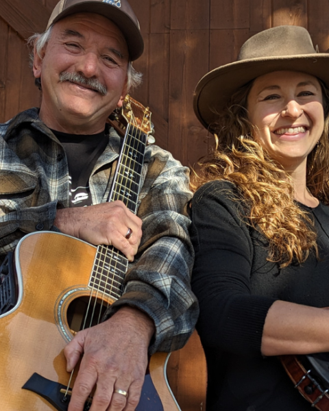 Lilli Worona & Mike Biggers are smiling while holding musical instruments—a guitar and a violin—against a wooden backdrop. Live music at Brunch at Riverhouse Lodge 8/10