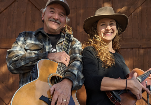 Lilli Worona & Mike Biggers are smiling while holding musical instruments—a guitar and a violin—against a wooden backdrop. Live music at Brunch at Riverhouse Lodge 8/10