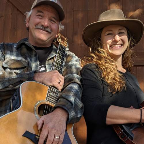 Lilli Worona & Mike Biggers are smiling while holding musical instruments—a guitar and a violin—against a wooden backdrop. Live music at Brunch at Riverhouse Lodge 8/10