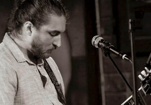 Kyle Hubbard is playing a guitar and standing in front of a microphone in a black and white image. Catch live show at Riverhouse Lodge in Bend Oregon 08/24