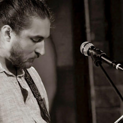 Kyle Hubbard is playing a guitar and standing in front of a microphone in a black and white image. Catch live show at Riverhouse Lodge in Bend Oregon 08/24