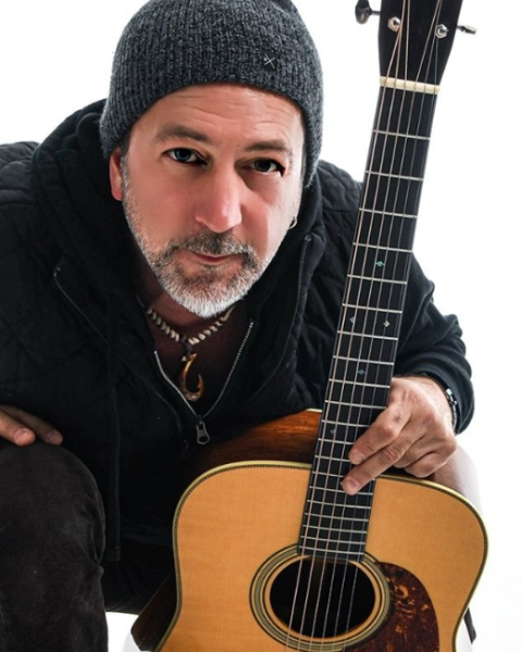 Pete Kartsounes wearing a beanie kneels, holding an acoustic guitar with a neutral background plays free live music at Riverhouse Lodge in Bend Oregon 08/21/25