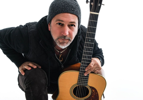 Pete Kartsounes wearing a beanie kneels, holding an acoustic guitar with a neutral background plays free live music at Riverhouse Lodge in Bend Oregon 08/21/25