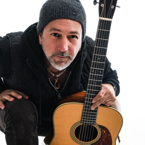 Pete Kartsounes wearing a beanie kneels, holding an acoustic guitar with a neutral background plays free live music at Riverhouse Lodge in Bend Oregon 08/21/25