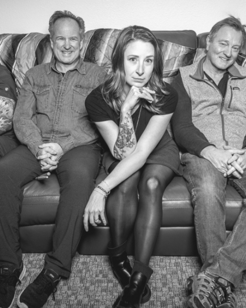 A black and white photo of the five member band Funk Around and Find Out sitting on a couch, with two holding guitars. They are smiling and look relaxed together. Funk Around and Find Out play Riverhouse Lodge in Bend Oregon 08/30/25 for free music