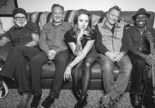 A black and white photo of the five member band Funk Around and Find Out sitting on a couch, with two holding guitars. They are smiling and look relaxed together. Funk Around and Find Out play Riverhouse Lodge in Bend Oregon 08/30/25 for free music