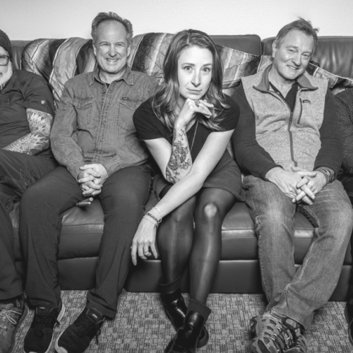 A black and white photo of the five member band Funk Around and Find Out sitting on a couch, with two holding guitars. They are smiling and look relaxed together. Funk Around and Find Out play Riverhouse Lodge in Bend Oregon 08/30/25 for free music