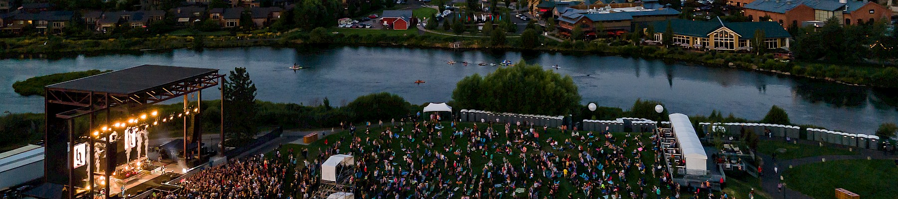 A large outdoor concert near a river, with a stage, audience, and tents, set against an evening cityscape and a distant hill.