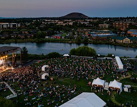 A large outdoor concert near a river, with a stage, audience, and tents, set against an evening cityscape and a distant hill.
