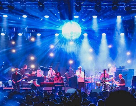 A live band performs on stage with bright lights and a disco ball, while a crowd watches. The atmosphere is vibrant and energetic.