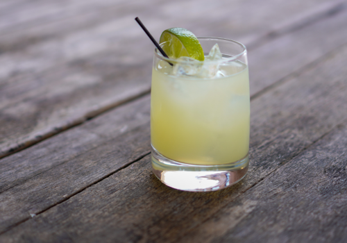 A glass of margarita with a lime wedge and straw, placed on a wooden surface. $7 House Margaritas at Currents at Riverhouse July 29. Margarita Monday awaits