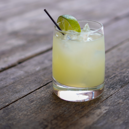 A glass of margarita with a lime wedge and straw, placed on a wooden surface. $7 House Margaritas at Currents at Riverhouse July 29. Margarita Monday awaits
