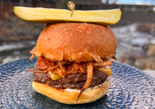 A burger with cheese and crispy onions, topped with a pickle slice on a blue plate. In the background, there's a river.