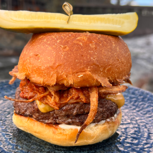 A burger with cheese and crispy onions, topped with a pickle slice on a blue plate. In the background, there's a river.