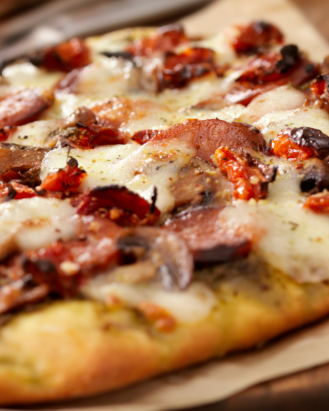 A close-up of a delicious flatbread pizza topped with sausage, mushrooms, cheese, and sun-dried tomatoes on parchment paper.